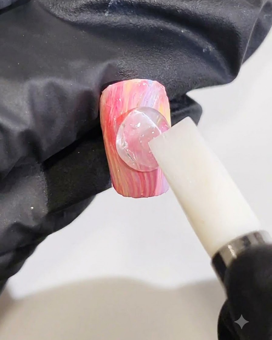 Una mano con guantes usando una herramienta de silicona blanca para dar forma a una gota transparente de gel 3D en una línea de onda sobre una uña de mármol rosa y amarilla.