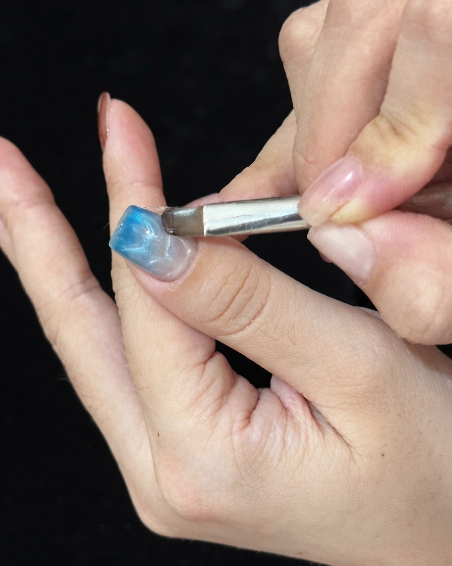 Un pincel aplicando gel de vidrio azul a toques en la punta de una uña manicurada para crear un degradado ombré.