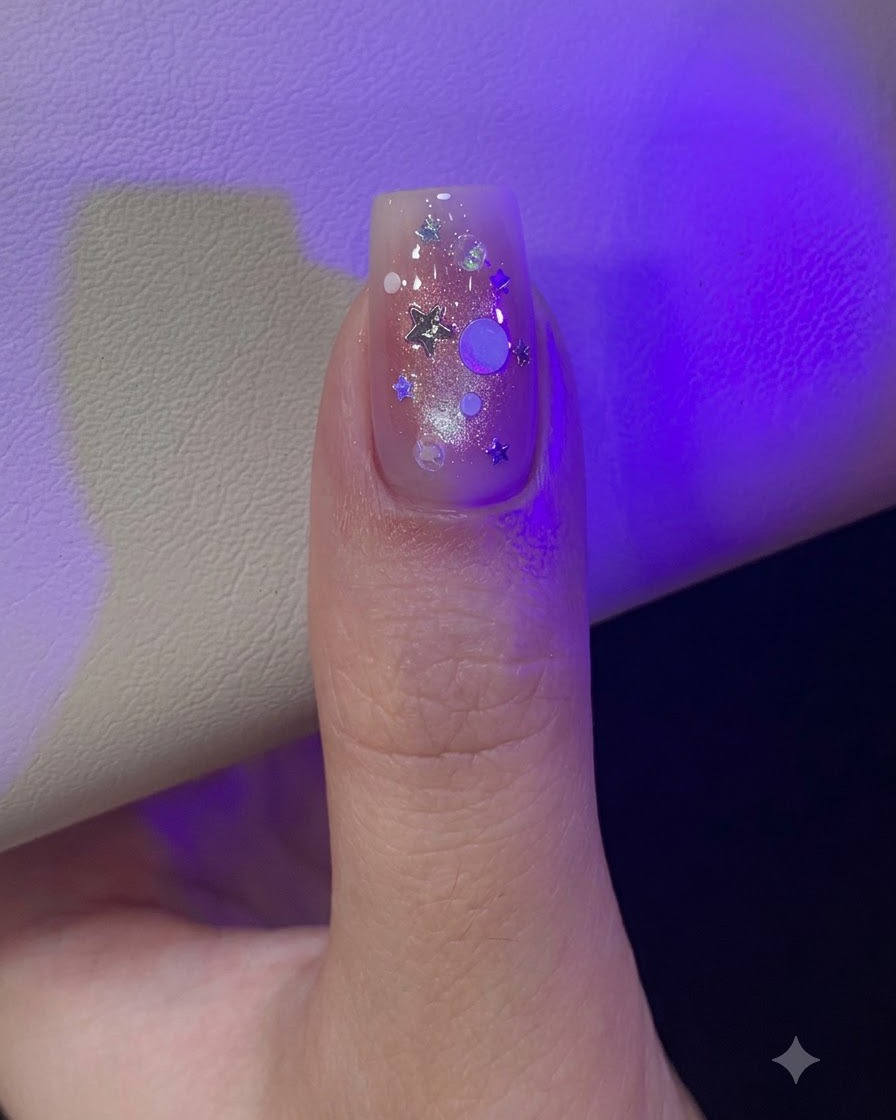 Un pulgar bajo la luz de la lámpara decorado con lentejuelas de glitter holográfico plateadas en forma de estrella y redondas dispersas.