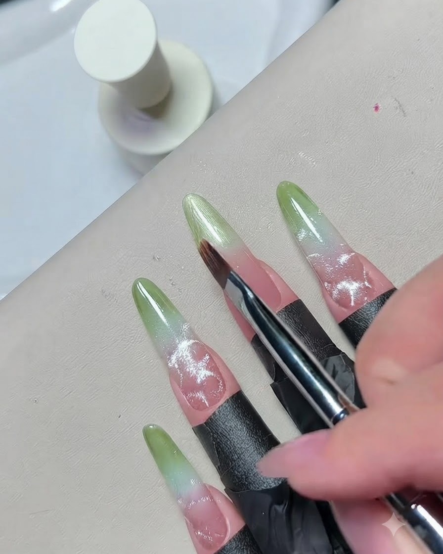 Un primer plano de un pincel fino difuminando el borde del esmalte en gel verde para crear una mezcla de degradado fluida.