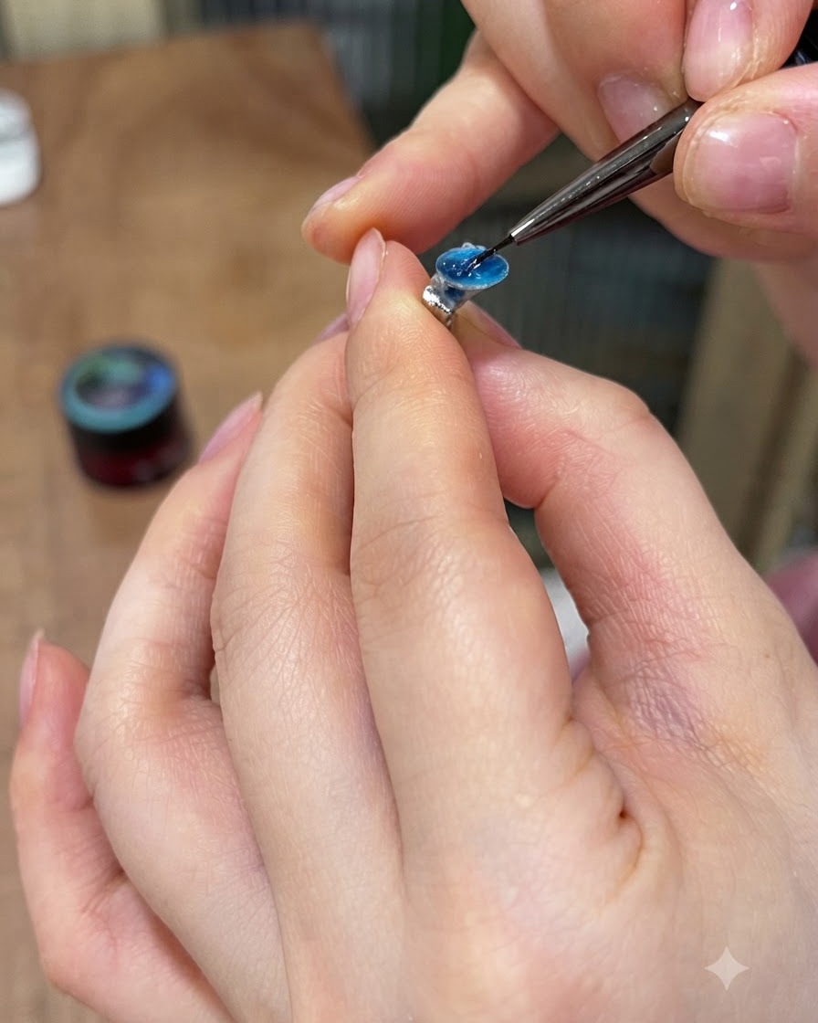 Un pincel de detalle aplicando pegamento sólido para pedrería en el reverso plano de una gema de gel azul.