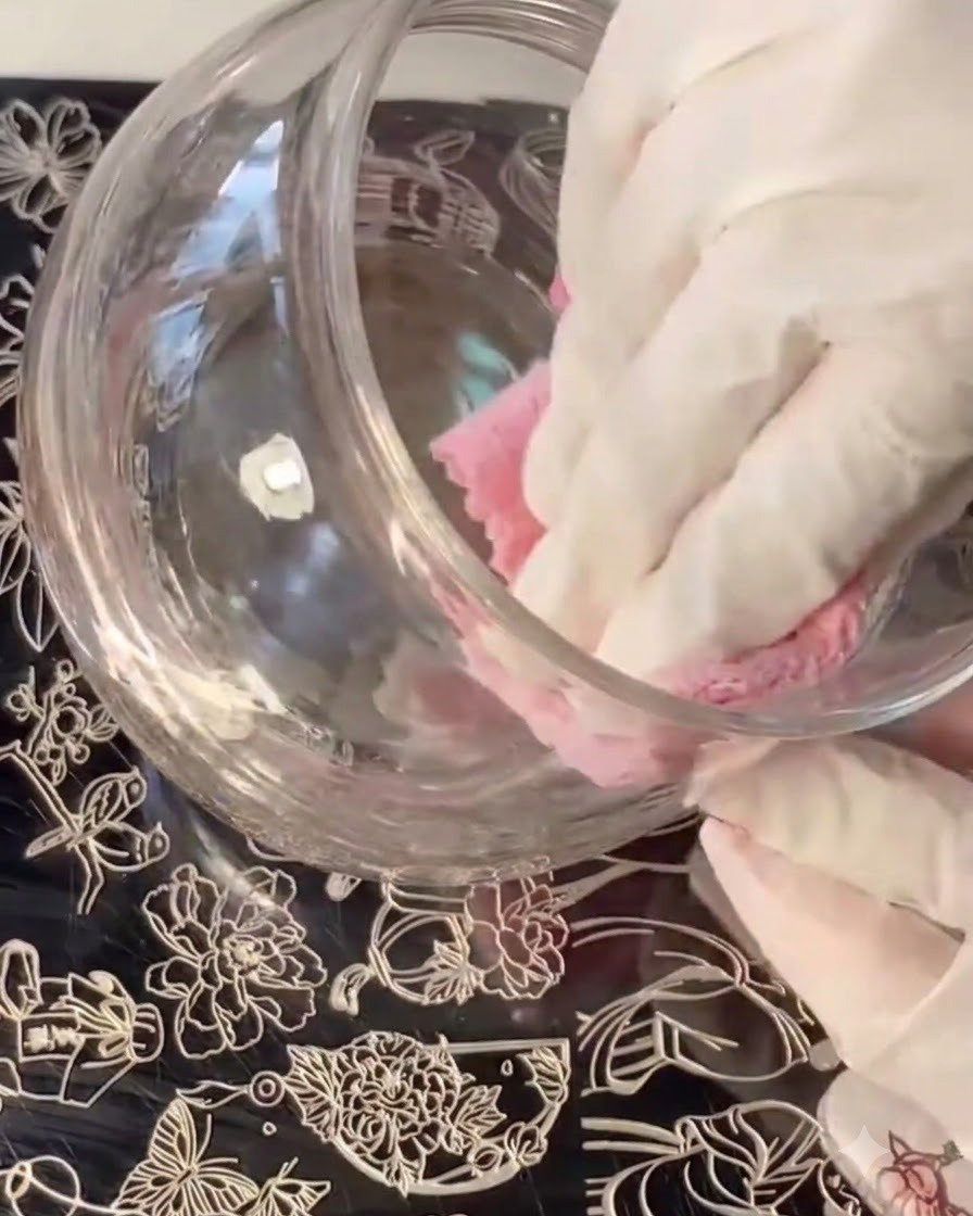 Una mano con guantes limpiando o pasando un paño por un recipiente de vidrio mientras placas de stamping con diseños florales yacen debajo.