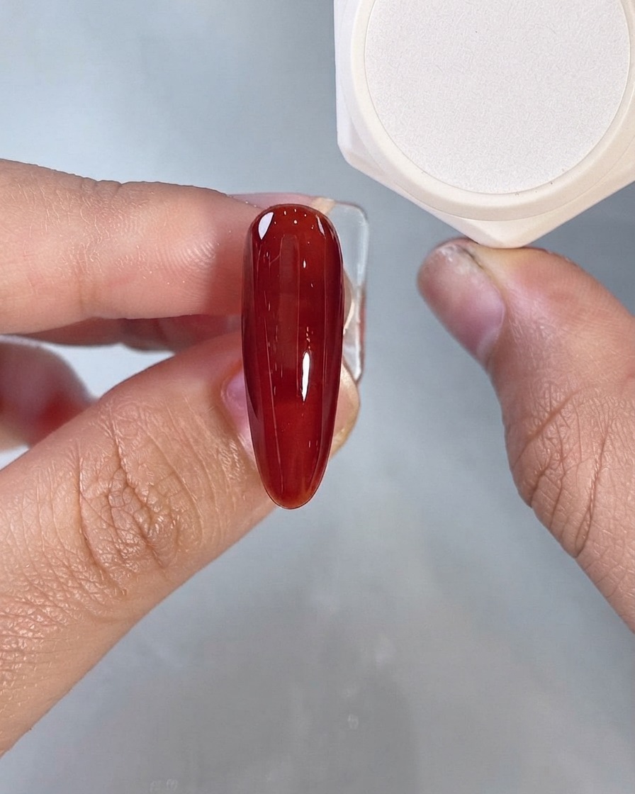 Un recubrimiento de gel líquido rojo oscuro cubriendo una sola uña postiza alargada de cerca.