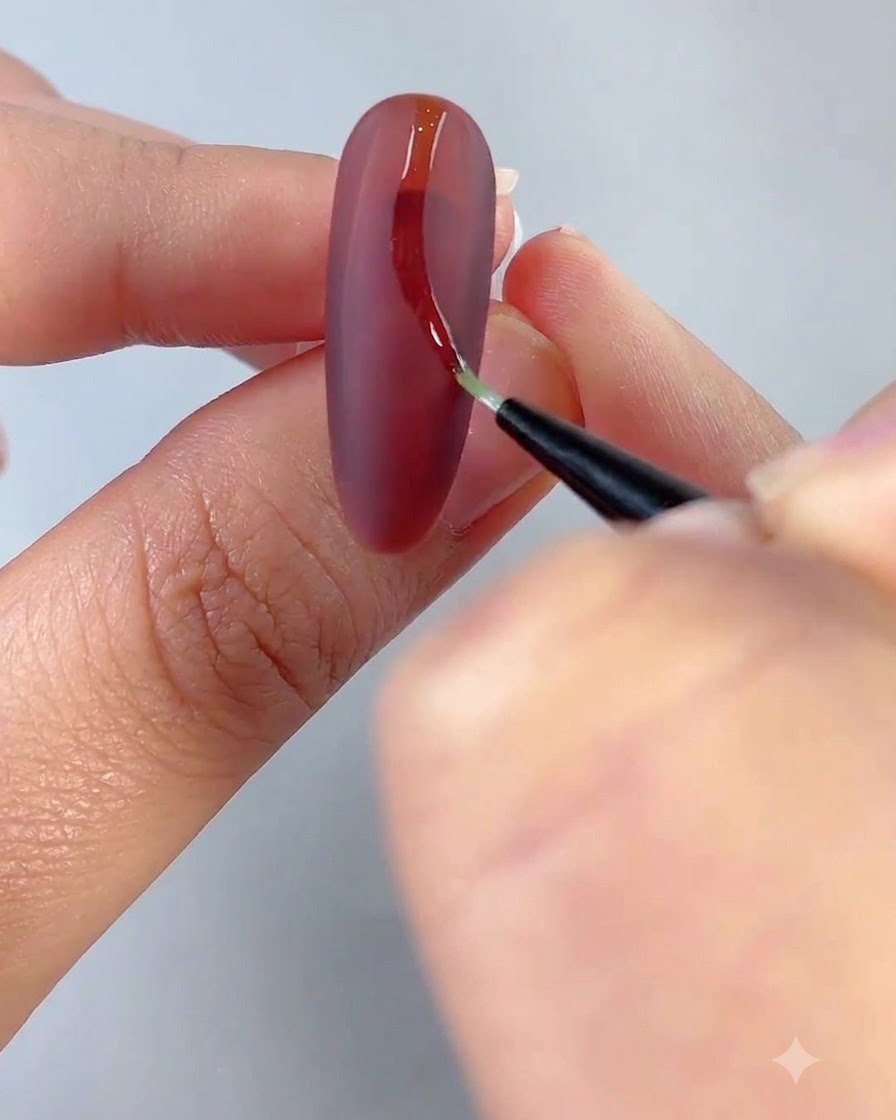 Un pincel fino de delineado dibujando una línea curva transparente sobre una superficie de uña roja mate.