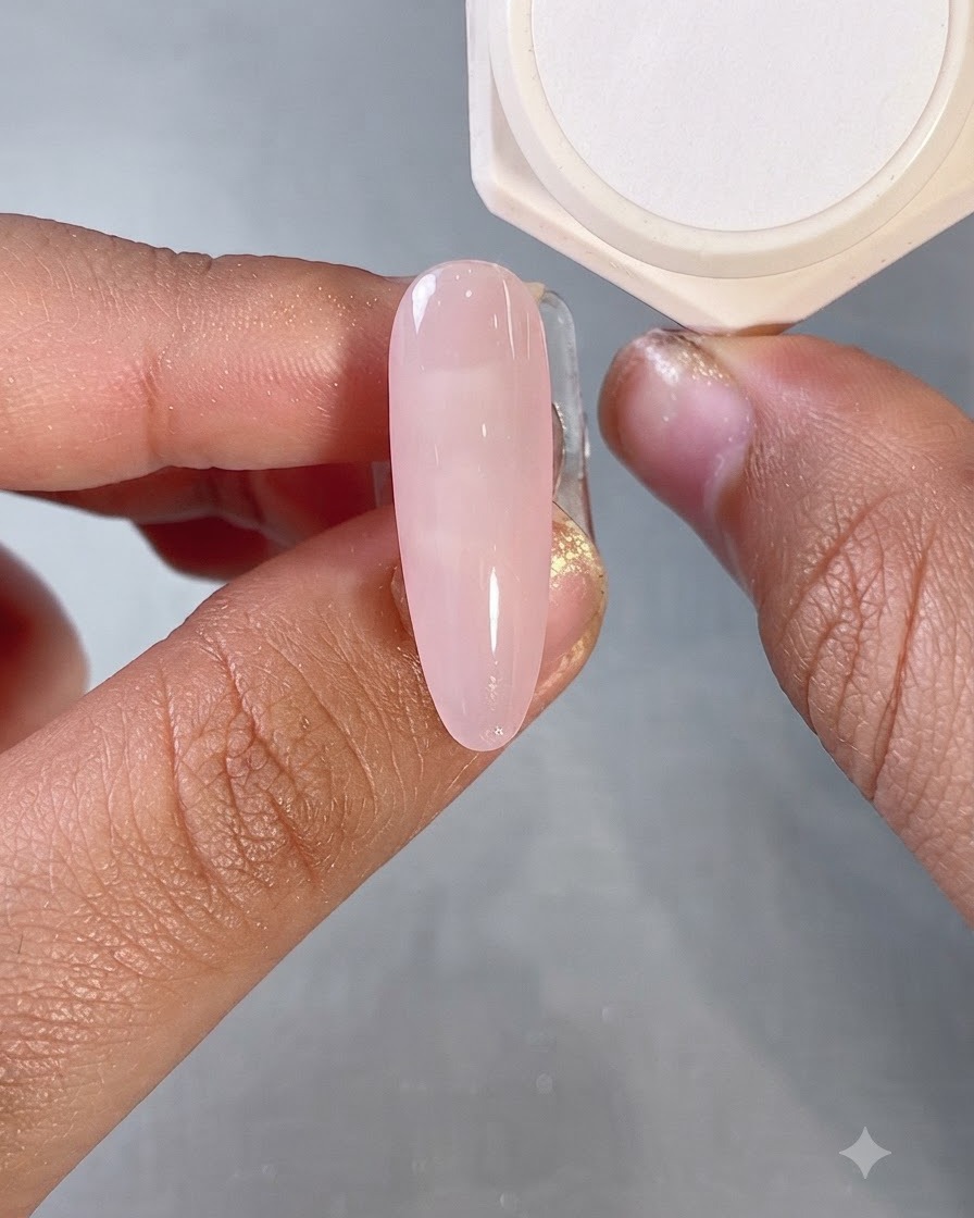 Un pincel aplicando una capa base de gel rosa lechoso translúcido sobre una uña de forma ovalada.