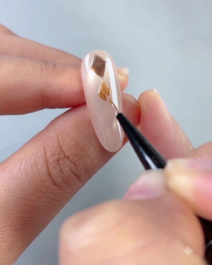 Un primer plano de un pincel de arte de uñas fino pintando una forma de diamante marrón en una uña rosa translúcida.