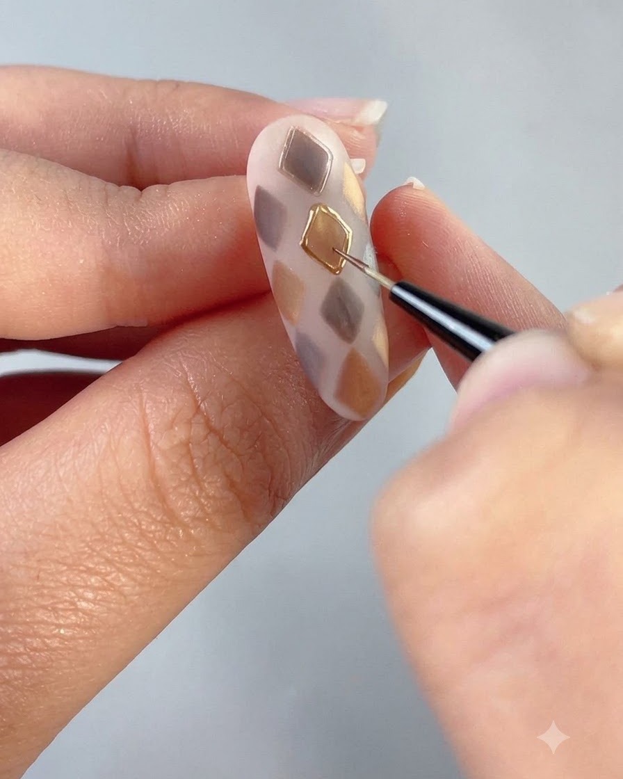Un pincel fino dibujando un contorno de gel transparente alrededor de una forma de diamante dorado en una uña decorada.