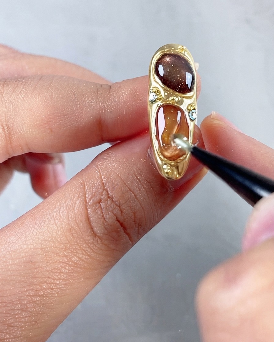 Un pincel fino alisando una capa superior brillante sobre un diseño de uñas ornamentado y multicolor con piedras preciosas y marco dorado.